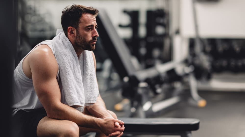 Mężczyzna po zakończonym treningu, z ręcznikiem na karku, odpoczywa siedząc na ławce w siłowni