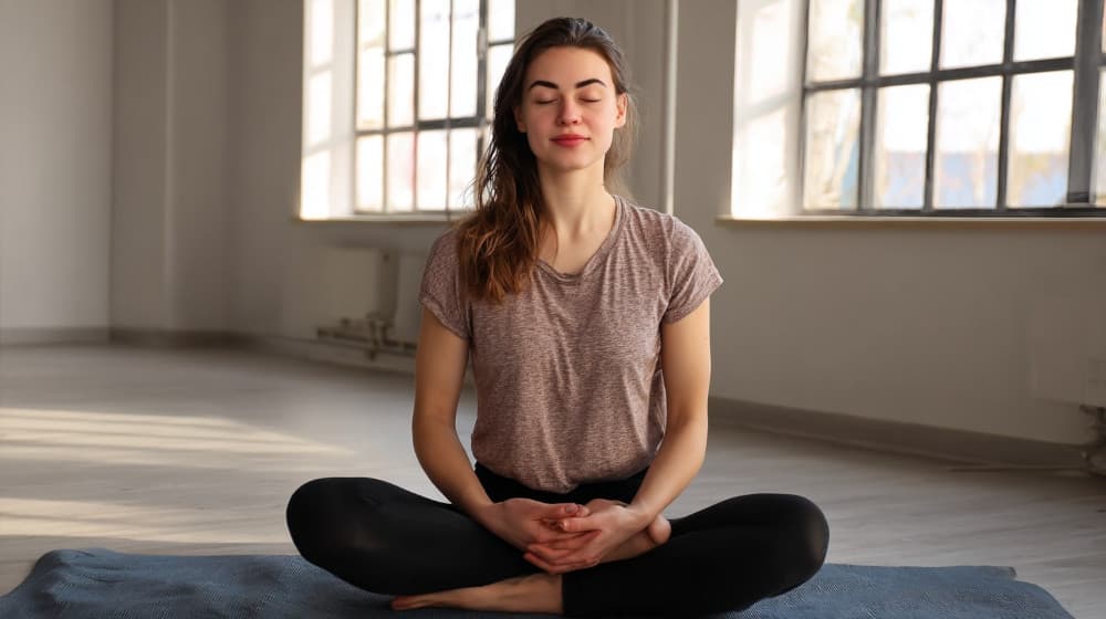 Młoda kobieta siedzi po turecku na macie, oczy zamknięte, spokojny oddech